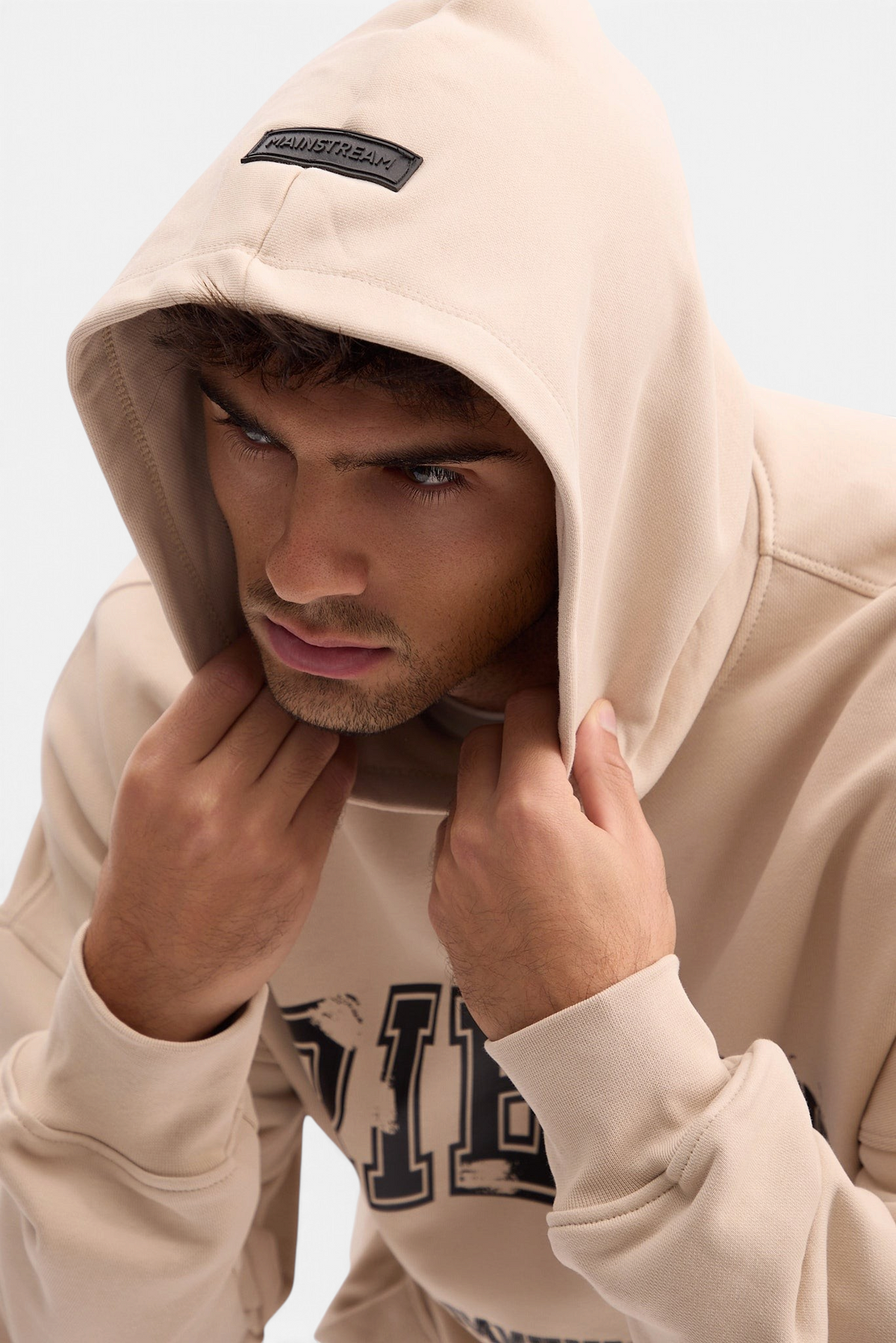 Mainstreamonline beige oversized hoodie with a hood and branded patch, worn by a man in a studio setting.