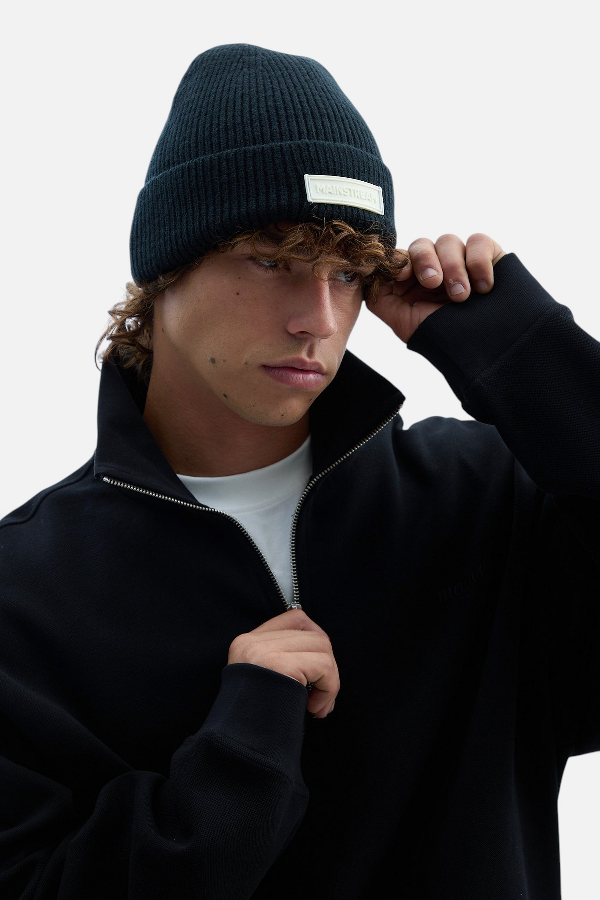 Mainstreamonline black ribbed knit beanie with a branded patch worn by a young man in a black zip-up sweatshirt