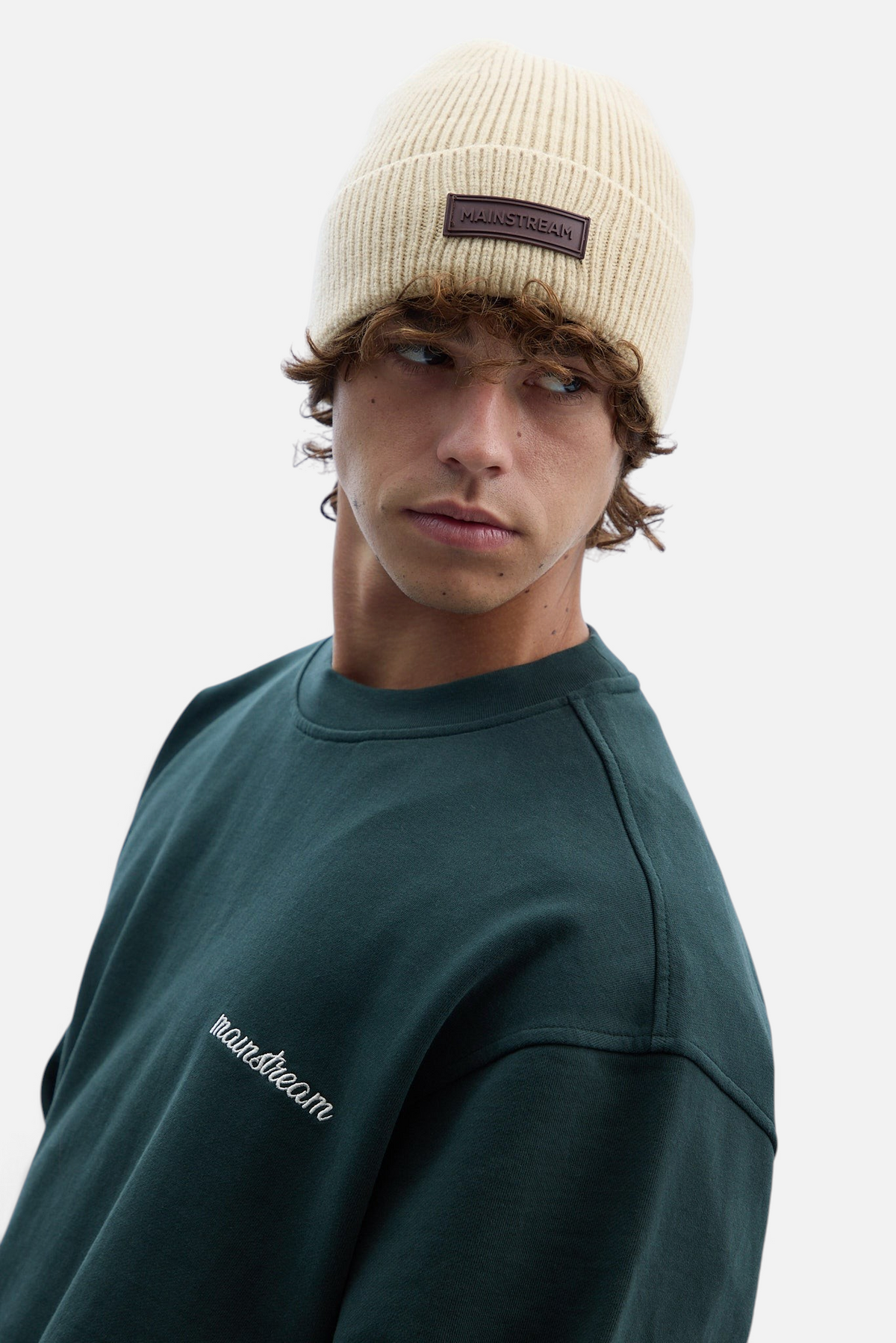 Mainstreamonline beige ribbed knit beanie with a brown leather logo patch worn by a man in a dark green sweatshirt